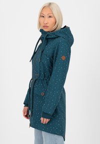 Parka impermeabile color teal con cappuccio regolabile, caratterizzata da un motivo a spruzzi multicolori, vita elastica e dettagli marroni sulle maniche.