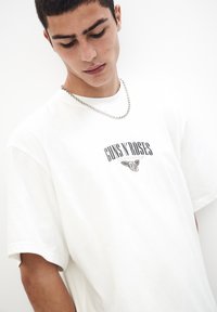 Hvid bomulds t-shirt med korte ærmer, der har et grafisk print af "GUNS N' ROSES" og et rosemblem. En sølv kædedesign er synligt.