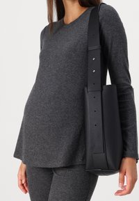 Svart läder axelväska med slät yta och justerbar rem, hålls av en person som bär en grå ribbad stickad topp och matchande byxor.