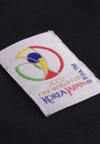 Wit stoffen patch met het logo van de FIFA Wereldbeker 2002 in rood, geel, groen en blauw, gestikt op een zwarte textielachtergrond.