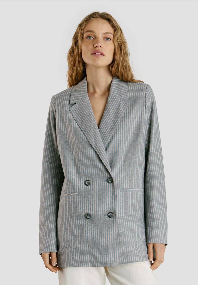 Kvinde iført lyseblå dobbeltradet blazer med tynde lodrette striber og hvide bukser, stående foran ensfarvet baggrund.