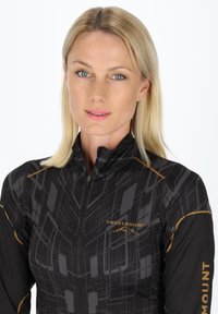 Top sportivo nero con zip, caratterizzato da pattern geometrici in grigio e oro, con il logo "SWEDEMOUNT" sul davanti. Design morbido e aderente.