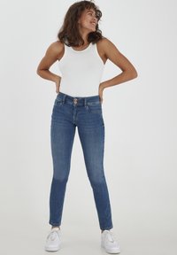 Jeans skinny de mezclilla azul con un lavado claro, cinco bolsillos, cierre de dos botones y un ligero estiramiento, combinados con una camiseta sin mangas blanca y zapatillas blancas.