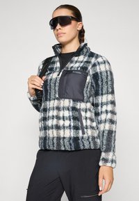 Fleecepullover i teal, hvit og grå rutete med halv-glidelås design; har en svart brystlomme og teksturert stoff.