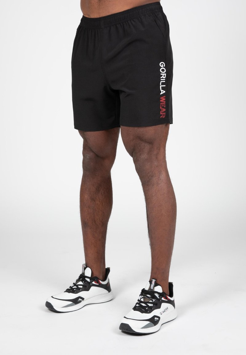 Zwarte sportieve shorts met een elastische tailleband, voorzien van witte en rode "GORILLA WEAR" tekst aan de linkerzijde, gecombineerd met zwart-witte sneakers.