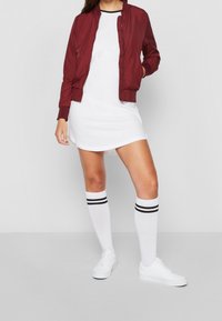 Burgunderfarbene Bomberjacke über einem weißen Kleid, kombiniert mit knielangen weißen Socken mit schwarzen Streifen und weißen Sneakers.