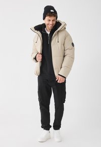 Jachetă puffer bej cu glugă, căptușeală din polar negru și patch cu logo. Purtați peste un hanorac negru și asortați cu pantalonii cargo și adidașii albi.