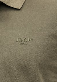 Κοντινό πλάνο μπεζ υφάσματος με ανάγλυφο λογότυπο «JOOP! JEANS» και ορατό διπλωμένο άκρο γιακά.