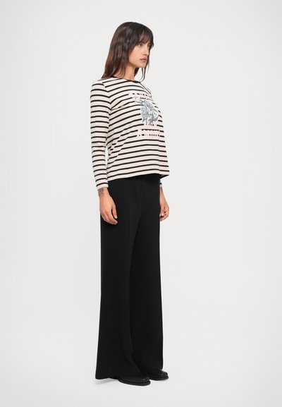 Striped long-sleeve shirt in beige and black with floral graphic, paired with black wide-leg trousers and flat shoes, shown from the side.