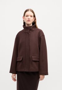 Hnědý vlněný kabát s vysokým límcem, volným střihem a dvěma předními kapsami. Hladká textura a minimalistický design, umístěný na jednobarevném pozadí.