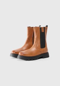 Botas de cuero marrón hasta el tobillo con paneles elásticos negros, suela de goma texturizada y un acabado suave, que presentan costuras mínimas y una lengüeta.