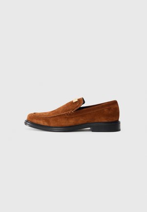 Brown suede loafers with a rounded toe, low black rubber sole, and decorative stitch detailing along the upper. Gold-tone accent on the side.