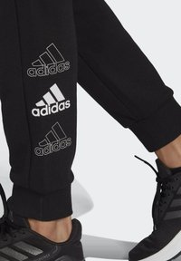 Svarta Adidas joggingbyxor har tre vita logotyp-accenter längs vänster ben. Tyget verkar mjukt med en manschettkant och en avsmalnande design.