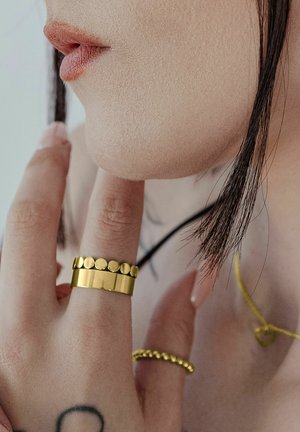 Nahaufnahme von Lippen, Kinn und Hand einer Person mit zwei goldenen Ringen, dazu eine goldene Halskette und eine Strähne dunkler Haare sichtbar.