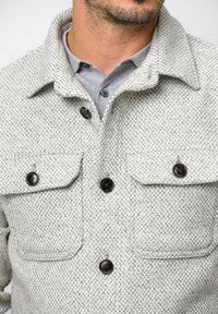 Graue strukturierte Jacke mit einem Kragen, die über zwei Brusttaschen und dunklen Knöpfen verfügt. Darunter ist ein graues Hemd mit Kragen sichtbar.