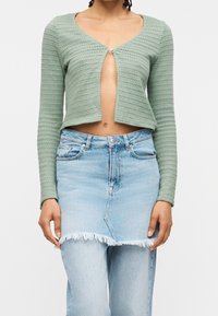 Personne portant un cardigan court en maille verte avec un bouton fermé et une jupe en jean bleu clair superposée à un jean.