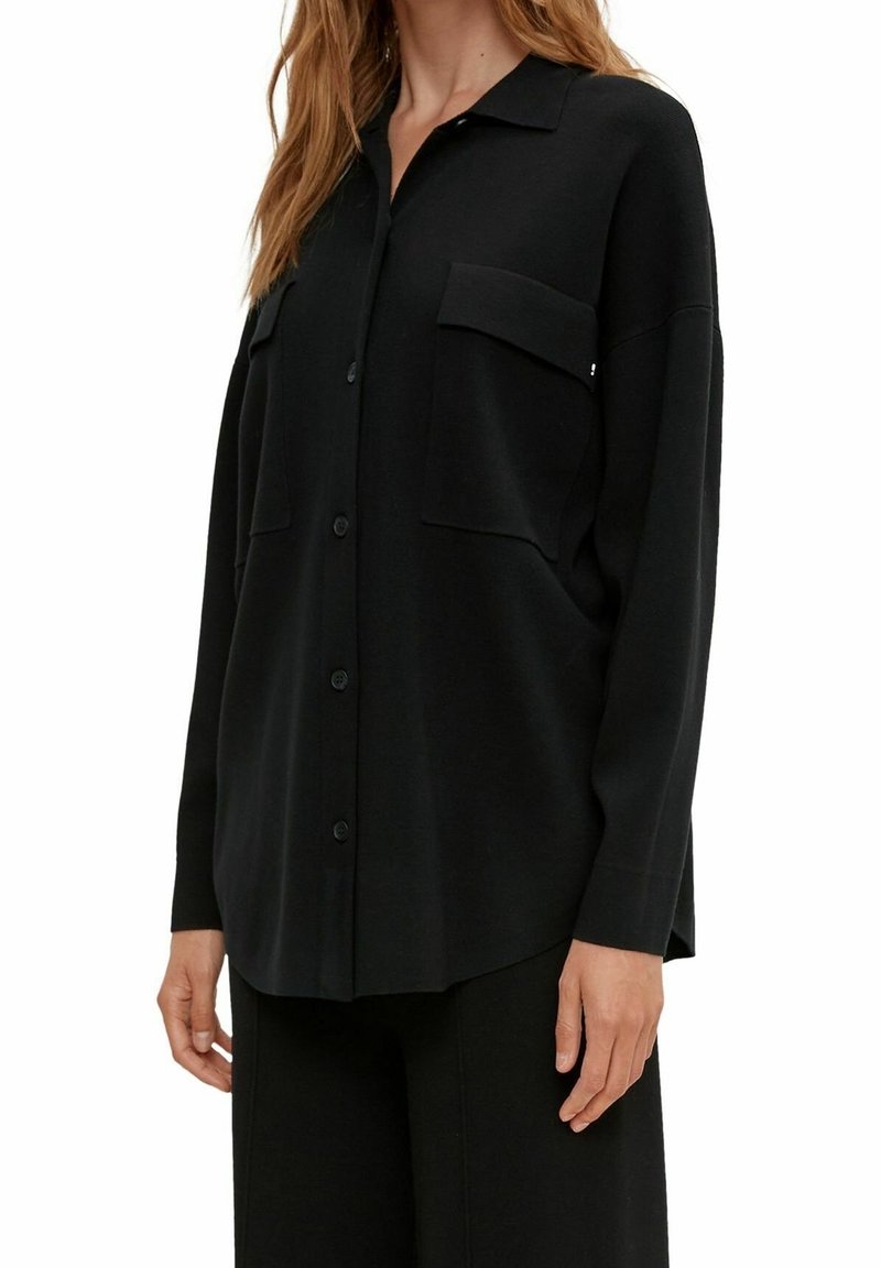 Femme portant une chemise noire ample à boutons avec poches poitrine et manches longues, associée à un pantalon noir.