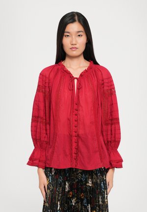Punainen blusa, jossa on rypytetty pääntie, napit edessä ja pitkät puffihihat, joissa on pitsikoristeet ja leveät hihansuut.