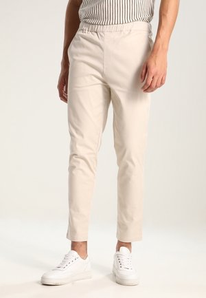 Mężczyzna ubrany w beżowe dopasowane spodnie, białe tenisówki i włożoną do środka czarno-białą koszulę w paski, stojący na jasnym tle.