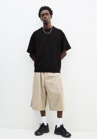 Černé oversized tričko s krátkým rukávem, béžové kraťasy do kolen, černé tenisky a bílé ponožky, na jednoduchém pozadí.