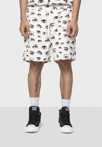Pantaloni corti bianchi con una stampa di occhi nera ripetuta, cintura elastica con cordino e tasche laterali, abbinati a sneakers nere alte.