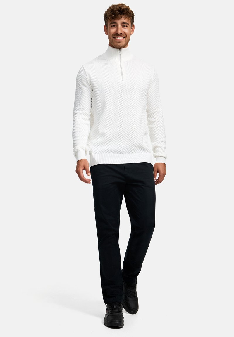 Suéter blanco con cremallera en el cuello, con un patrón de espiga texturizado, combinado con pantalones negros y zapatos negros, modelo caminando frente a un fondo liso.