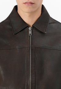 Homme portant une veste en cuir marron foncé avec zip et large col, gros plan sur la poitrine et le bas du visage sur fond blanc.