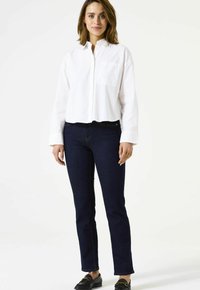Chemise blanche à boutons avec une poche poitrine, associée à un jean bleu foncé. La tenue présente une coupe décontractée et un design minimaliste.