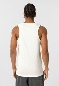 Witte geribde tanktop met een ronde halslijn en brede armsgaten, gedragen door een persoon die zijn of haar rug toont, met gevlochten haar en grijze broek.