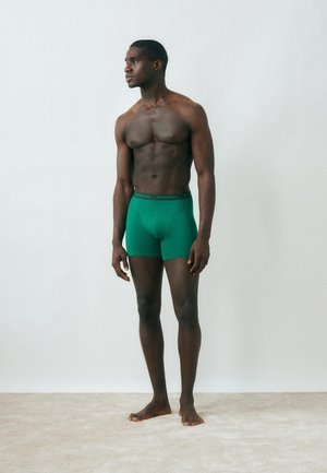 Jeune homme noir debout pieds nus sur un tapis, portant un boxer vert, regardant sur le côté contre un mur blanc uni.