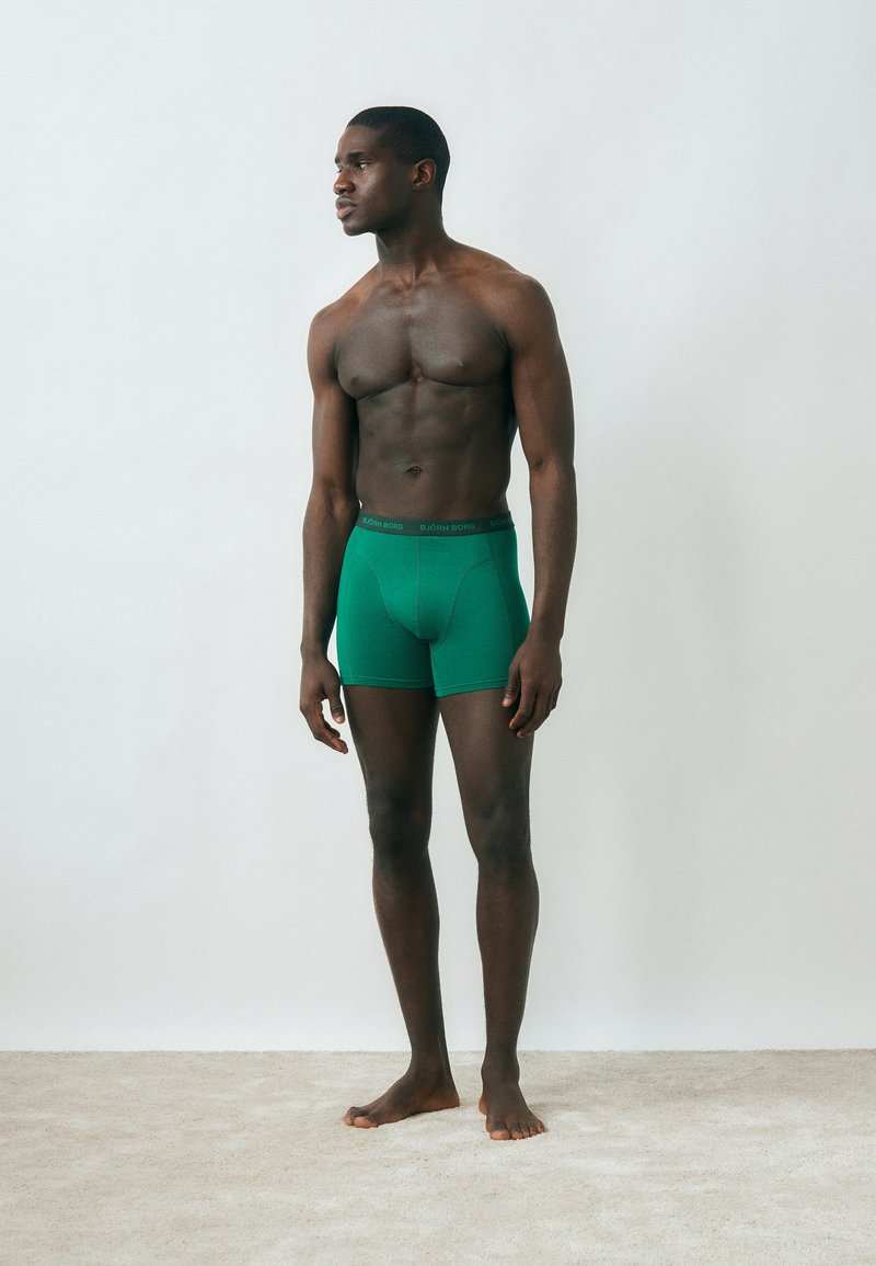 Jeune homme noir debout pieds nus sur un tapis, portant un boxer vert, regardant sur le côté contre un mur blanc uni.
