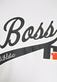 Valkoinen lyhytnauhainen t-paita, jossa on musta ja punainen "Boss" -grafiikka ja "Athletic" -teksti kaarevassa muodossa edessä.