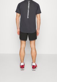 Camisa negra de manga corta con el logo blanco de "Under Armour" en la parte posterior, combinada con pantalones cortos negros y zapatillas deportivas rojas. Textura suave.