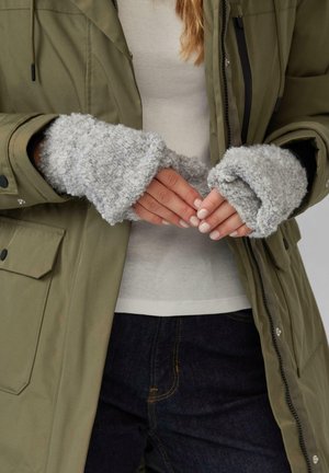 Graue fingerlose Handschuhe aus weichem, strukturiertem Material, getragen mit einem olivgrünen Mantel über einem hellen Shirt, die Hände überkreuzt.