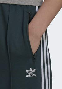 Mörkgrön adidas sweatpants med vita ränder, dragkedjefickor på sidorna och en liten logotyp på vänster ben. Slät tygkvalitet.