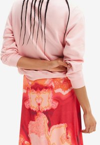 Hellrosa Sweatshirt mit gerippten Bündchen und lockerem Schnitt, kombiniert mit einem lebhaft rot-orange gemusterten Rock mit abstrakten Designs.