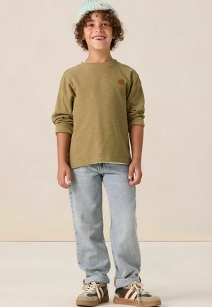 Chemise à manches longues côtelée olive avec un petit logo, jeans droits bleu clair, et baskets beiges avec des accents rouges et verts.