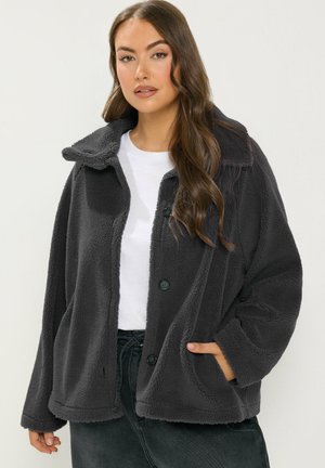 Giovane donna con lunghi capelli castani che indossa una giacca in pile grigio scuro con bottoni sopra una camicia bianca, con una mano nella tasca della giacca.