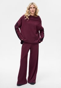 Bordeauxrode trui en wijde broekset, met zwarte gestructureerde accenten op de mouwen. Zachte stof met een ontspannen pasvorm en een gedrapeerde halslijn.