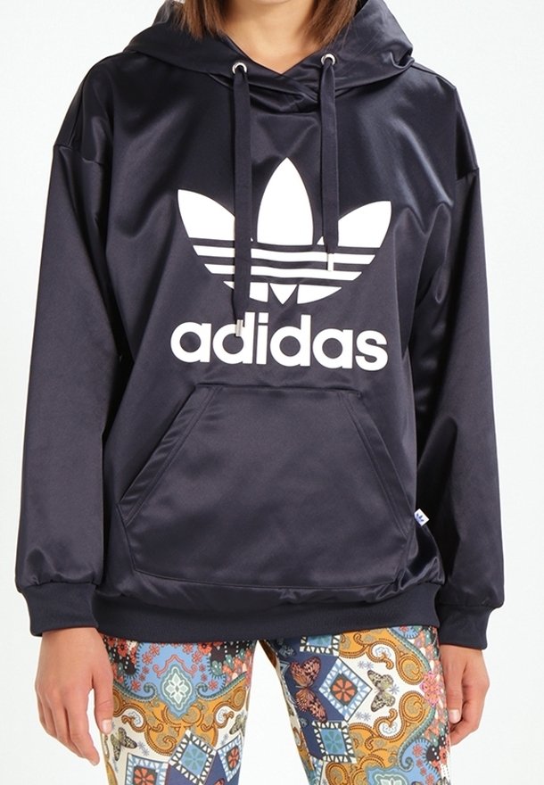 Marinblå satinhoodie med en vit Adidas-logotyp och framficka. Har en dragsko i huven och lös passform. Kombineras med mönstrade leggings.