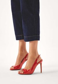 Punaiset lakatut nahkaiset slingback-kengät avoimilla varpailla ja ohuella korkokengällä, joissa on säädettävä nilkkaremmi ja kiiltävä pinta.