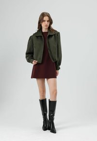 Veste courte vert olive avec manches bouffantes, associée à une robe bordeaux et des bottes noires montant jusqu'aux genoux. Tissu lisse et accessoires minimaux.