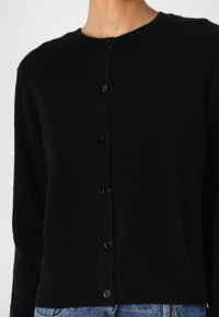 Svart strikket cardigan med rund hals, med syv svarte knapper foran og en myk, litt fuzzy tekstur.