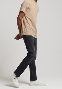 Camiseta de algodón beige con cuello redondo y un bolsillo en el pecho, combinada con unos vaqueros gris oscuro. Zapatillas blancas completan el look casual.