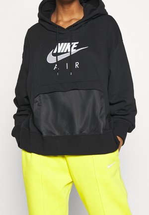 Czarna bluza Nike z szarym siatkowym panelem, regulowanym kapturem ze sznurkiem i białym nadrukiem logo. W połączeniu z jasnymi żółtymi spodniami.