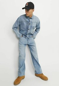 Veste en denim bleu clair et jeans, avec une casquette noire ajustée arborant un logo, et des bottes marron à lacets, créant une tenue décontractée.