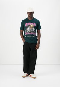 Green cotton t-shirt with "ADIDAS ORIGINALS" graphic in pink and yellow, paired with black wide-leg pants and white sneakers with red stripes.