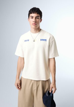 Mladý muž v krémovej košeli s krátkym rukávom s modrým textom a béžovými nohavicami drží námornícku modrú čiapku na svetlo šedom pozadí.