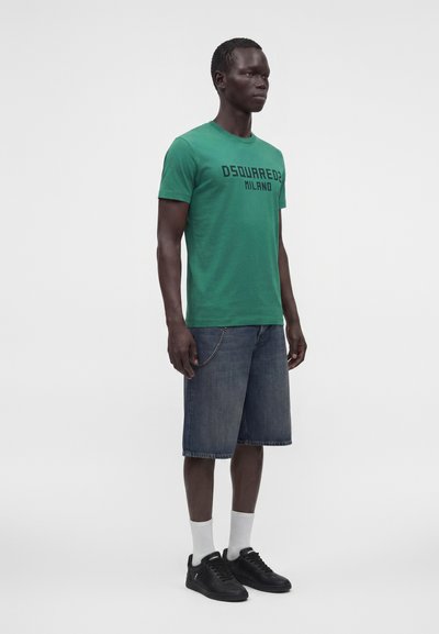 Άνδρας με πράσινο T-shirt DSQUARED2 Milano, σκουρόχρωμα τζιν σορτς μέχρι το γόνατο, λευκές κάλτσες και μαύρα αθλητικά παπούτσια σε απλό φόντο.