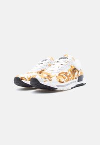 Άσπρα sneakers με χρυσά μπαρόκ σχέδια, μαύρες λαστιχένιες σόλες, άσπρα κορδόνια και την επωνυμία «Versace Jeans Couture» στο πλάι και στη γλώσσα.